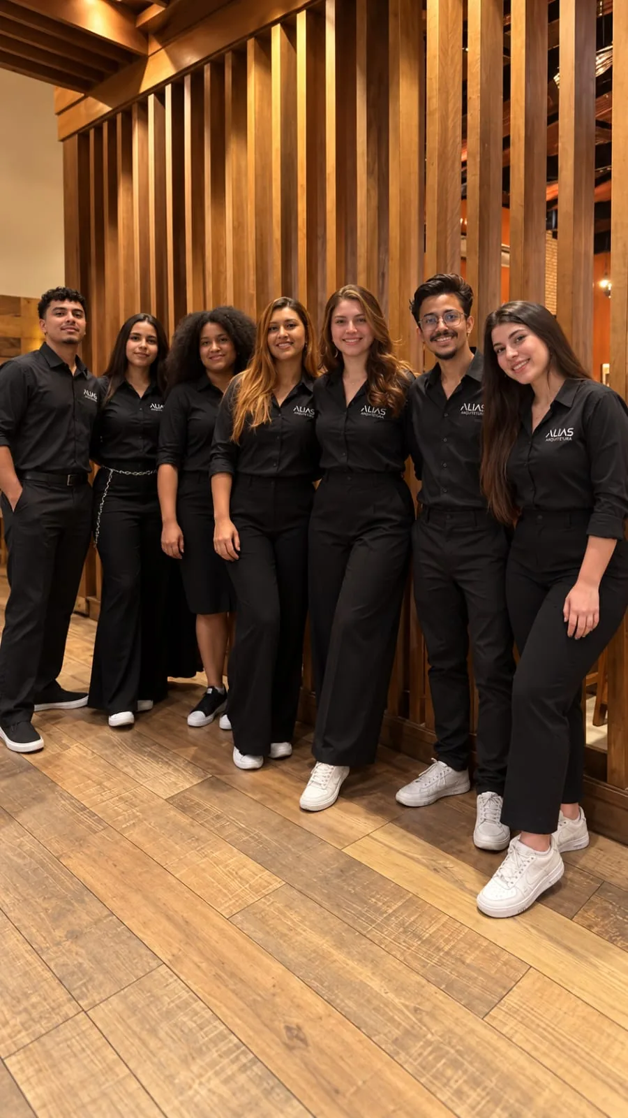 Time da Alias Arquitetura: sete pessoas de camisa preta com o logotipo da marca, lado a lado diante de um painel ripado de madeira