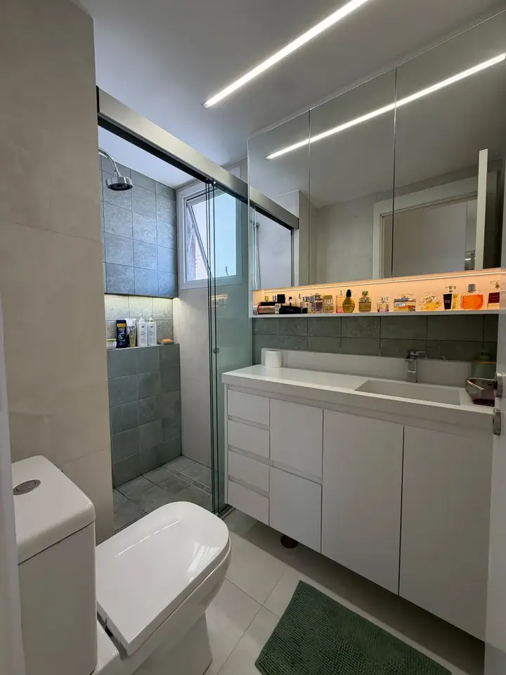 Banheiro com bancada branca, cuba esculpida, nicho iluminado, box de vidro e revestimento cinza