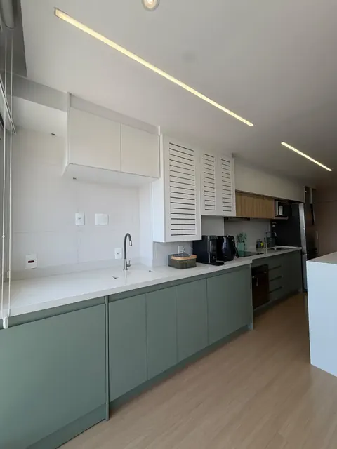 Cozinha com armários verdes, bancada clara, armários altos brancos com venezianas e iluminação linear no forro
