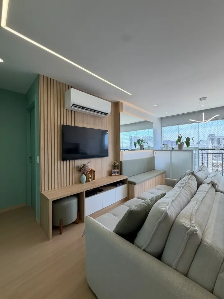 Sala de estar com painel ripado de madeira, TV, sofá claro, parede verde e varanda envidraçada ao fundo